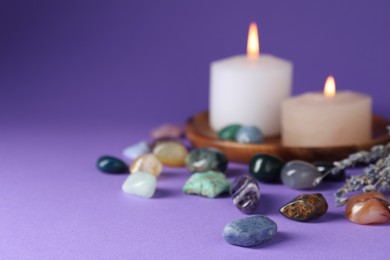 Photo of Different beautiful gemstones, lavender flowers and burning candles on purple background, closeup. Space for text