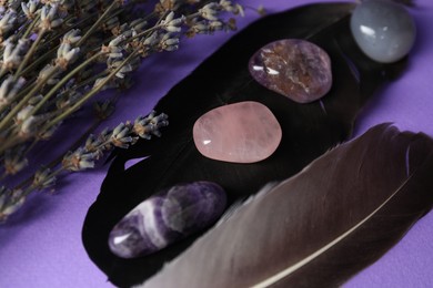 Photo of Different beautiful gemstones in bowl, feathers and lavender flowers on purple background, closeup