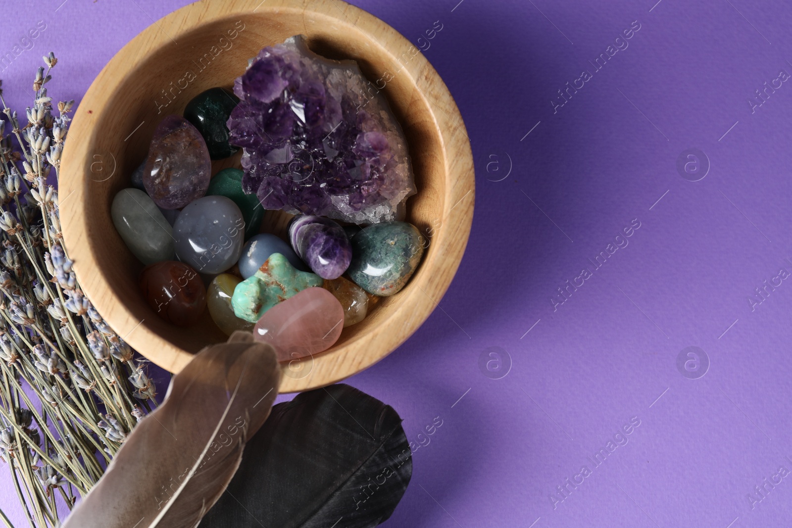 Photo of Different beautiful gemstones in bowl, feathers and lavender flowers on purple background, flat lay. Space for text