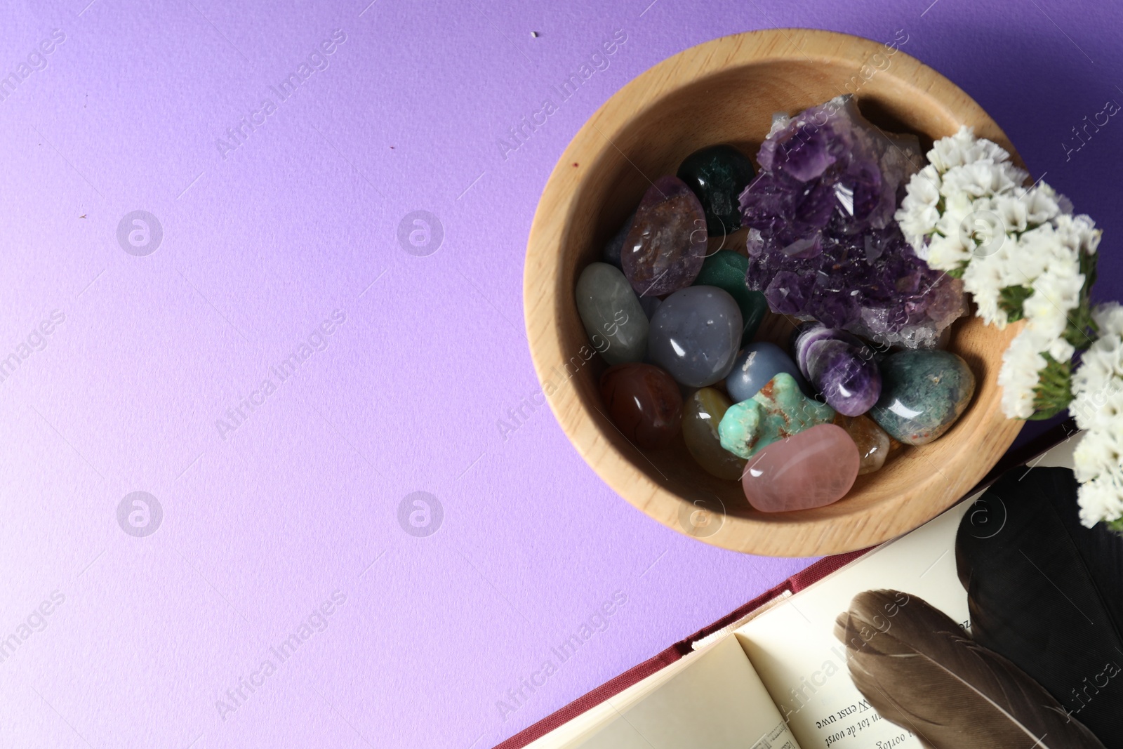 Photo of Different beautiful gemstones, feathers, flowers and book on purple background, flat lay. Space for text