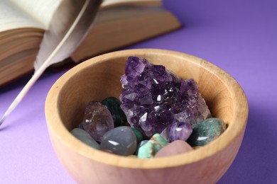 Photo of Different beautiful gemstones in bowl, feather and book on purple background, closeup