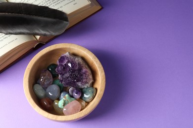 Photo of Different beautiful gemstones in bowl, feather and book on purple background, flat lay. Space for text