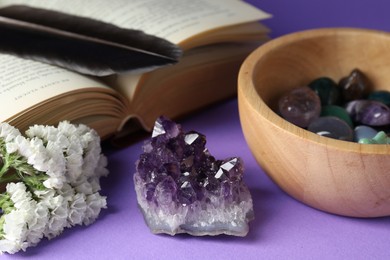 Photo of Different beautiful gemstones, feather, flowers and book on purple background, closeup
