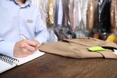Photo of Dry-cleaning service. Worker with jacket writing in notebook at counter indoors, closeup