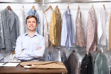 Photo of Dry-cleaning service. Happy worker at counter near rack with clothes in plastic bags indoors, space for text