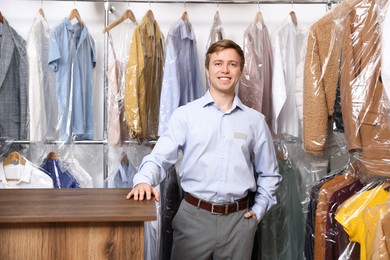Photo of Dry-cleaning service. Happy worker at counter near rack with clothes in plastic bags indoors