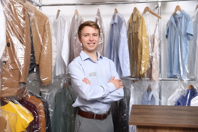 Photo of Dry-cleaning service. Happy worker near rack with clothes in plastic bags indoors