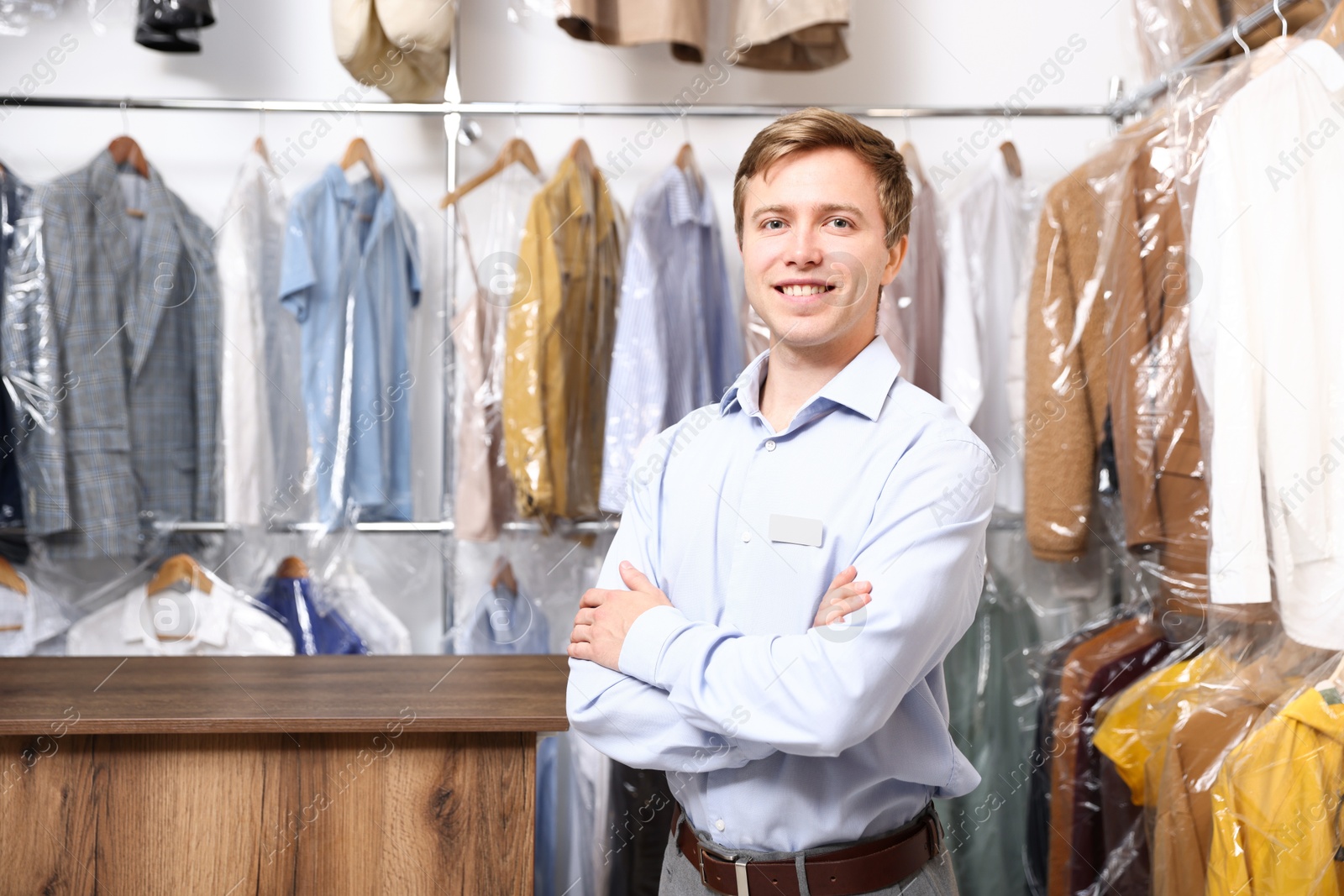 Photo of Dry-cleaning service. Happy worker near rack with clothes in plastic bags indoors, space for text