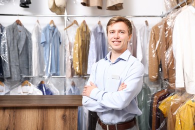 Photo of Dry-cleaning service. Happy worker near rack with clothes in plastic bags indoors, space for text