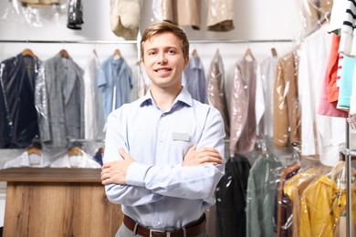 Photo of Dry-cleaning service. Happy worker near rack with clothes in plastic bags indoors