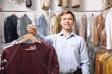 Photo of Dry-cleaning service. Happy worker holding jacket in plastic bag indoors