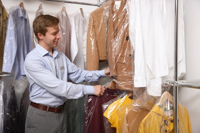 Photo of Dry-cleaning service. Happy worker choosing clothes from rack indoors