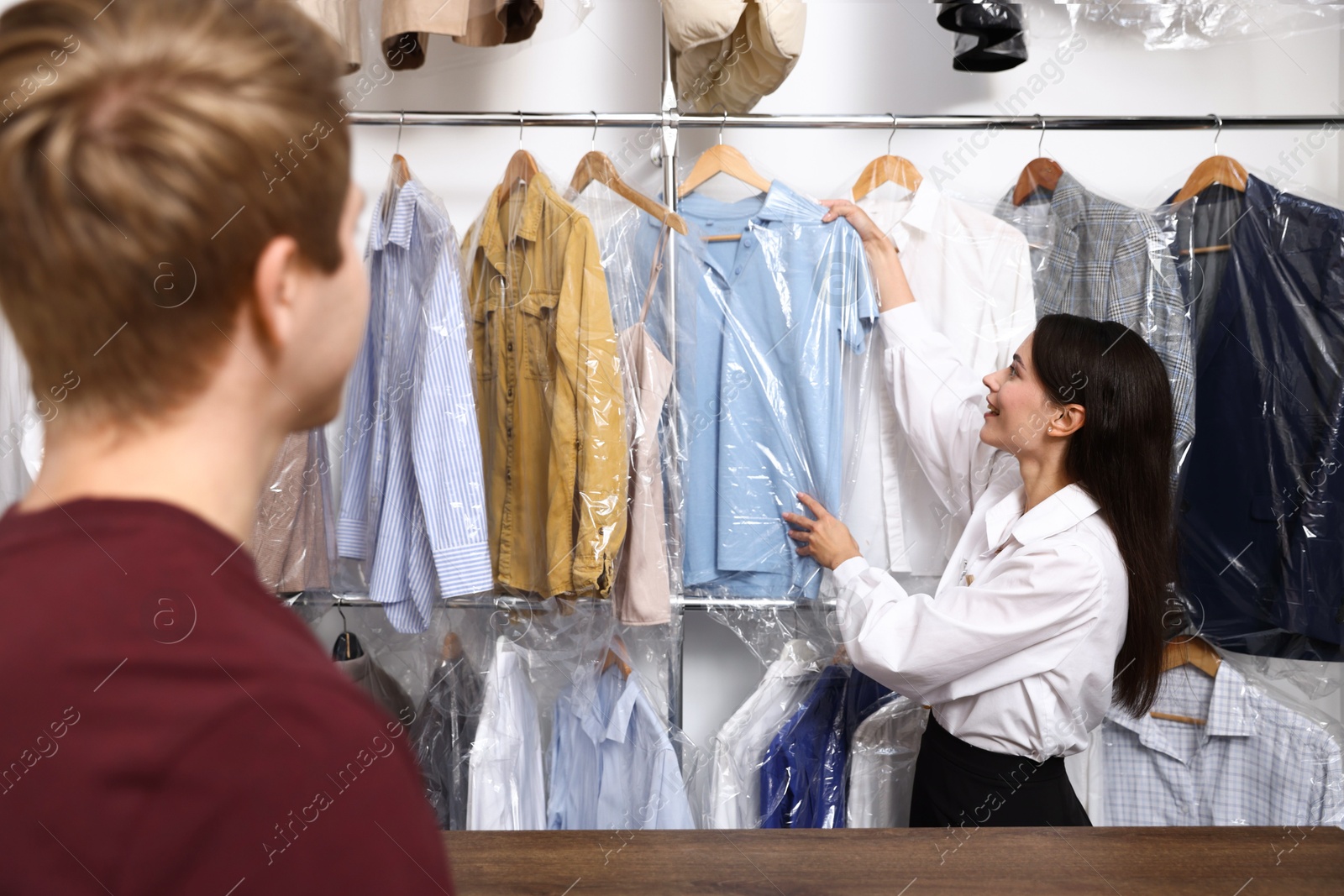 Photo of Dry-cleaning service. Happy worker choosing clothes from rack for client indoors