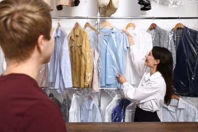 Photo of Dry-cleaning service. Happy worker choosing clothes from rack for client indoors