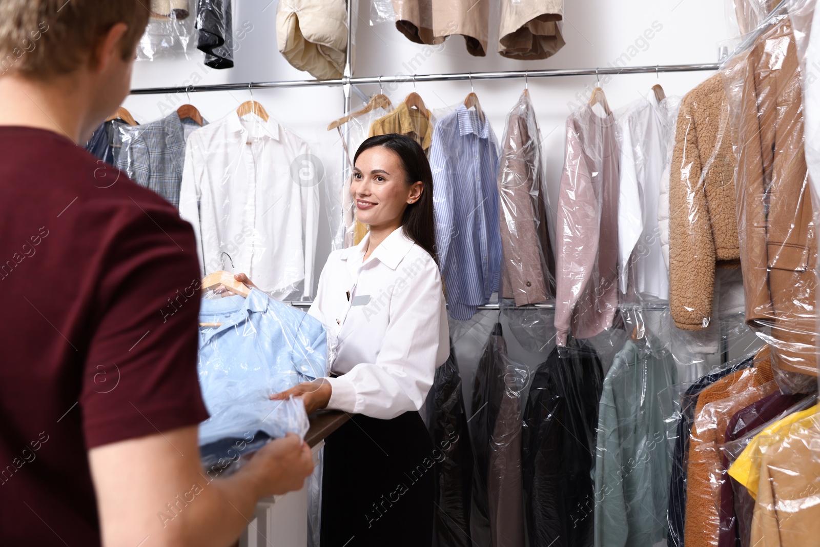 Photo of Dry-cleaning service. Happy worker giving shirt in plastic bag to client at counter indoors