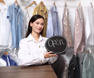 Photo of Dry-cleaning service. Happy worker with Open sign at counter indoors