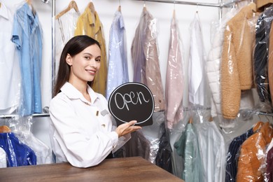 Photo of Dry-cleaning service. Happy worker with Open sign at counter indoors