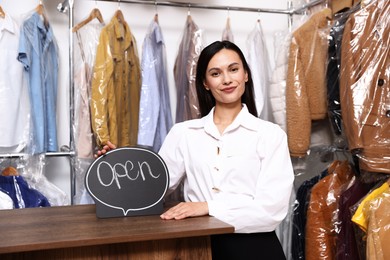 Photo of Dry-cleaning service. Happy worker with Open sign at counter indoors
