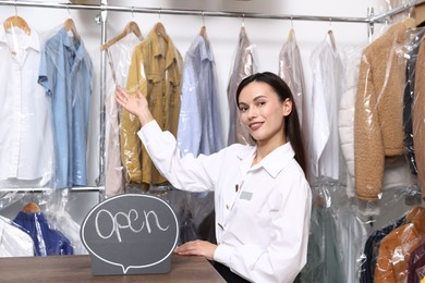 Photo of Dry-cleaning service. Happy worker with Open sign at counter indoors