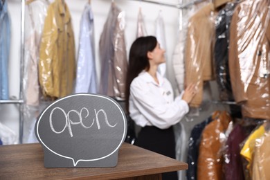 Photo of Dry-cleaning service. Worker choosing clothes from rack, focus on Open sign