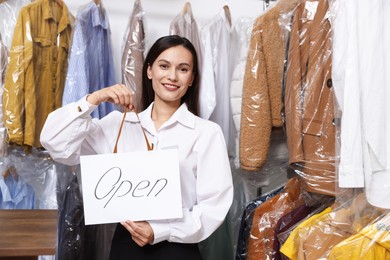 Photo of Dry-cleaning service. Happy worker holding Open sign at near rack with clothes indoors
