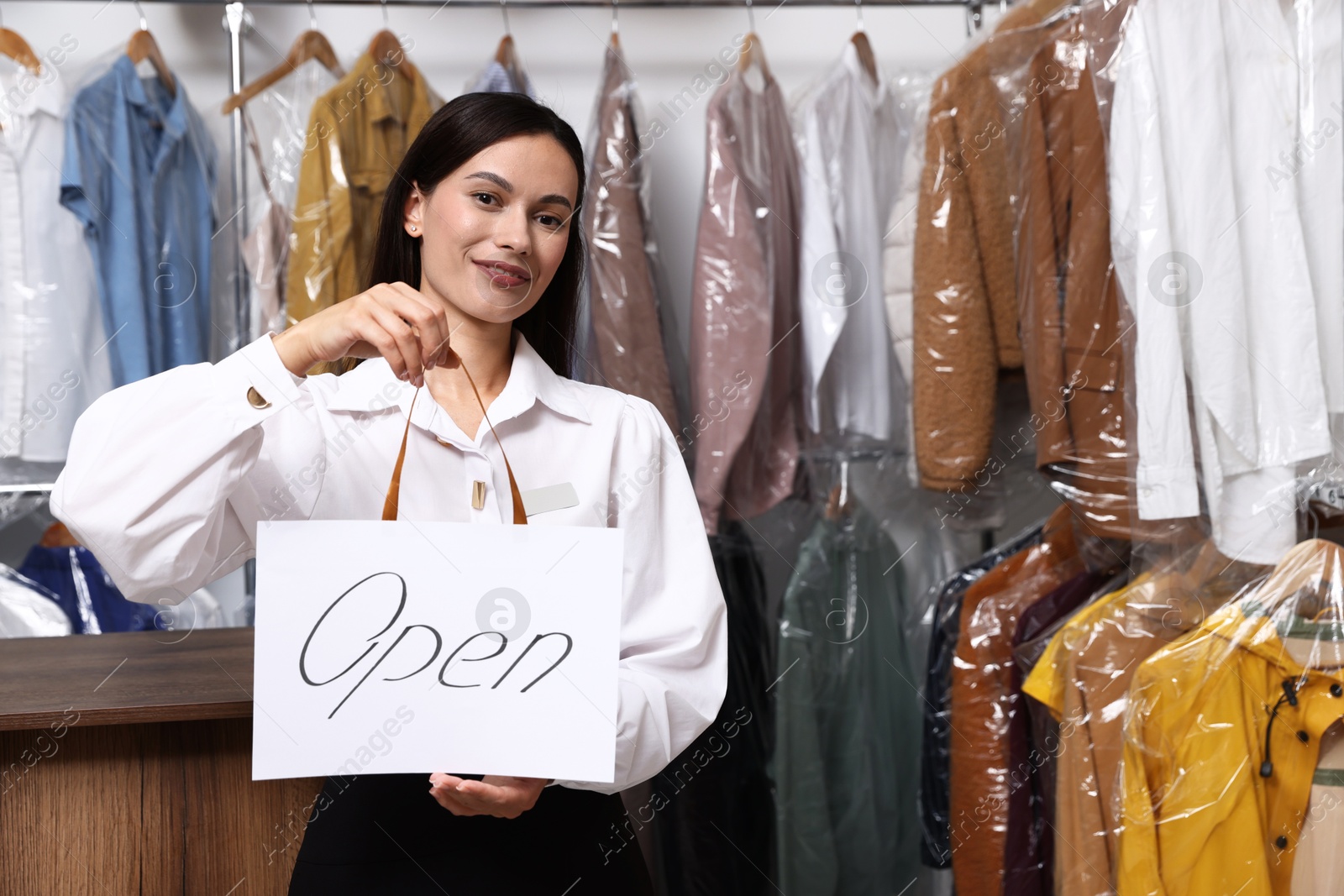 Photo of Dry-cleaning service. Happy worker holding Open sign at near rack with clothes indoors