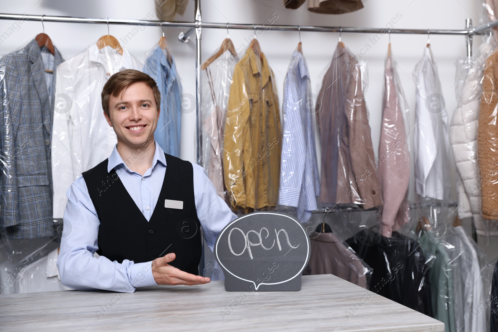Photo of Dry-cleaning service. Happy worker with Open sign at counter indoors