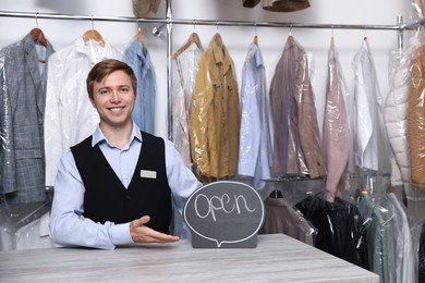 Photo of Dry-cleaning service. Happy worker with Open sign at counter indoors