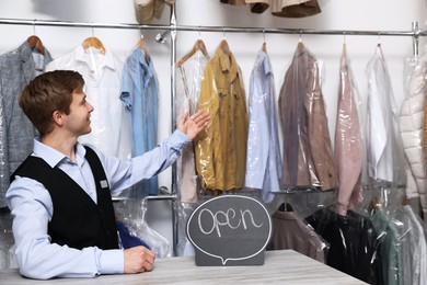 Photo of Dry-cleaning service. Happy worker with Open sign at counter indoors