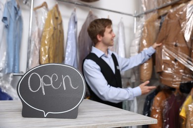 Photo of Dry-cleaning service. Worker choosing clothes from rack, focus on Open sign