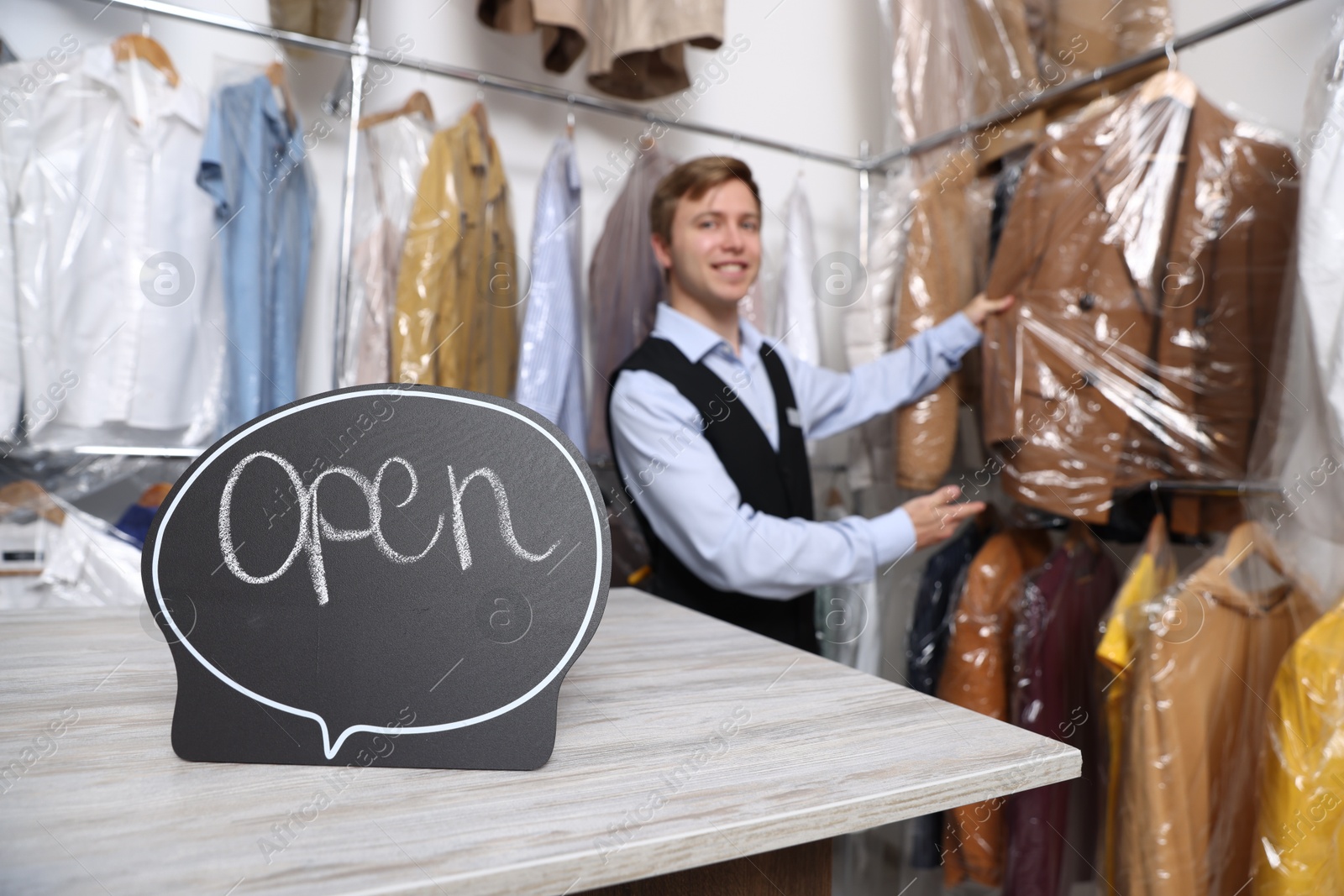 Photo of Dry-cleaning service. Worker choosing clothes from rack, focus on Open sign