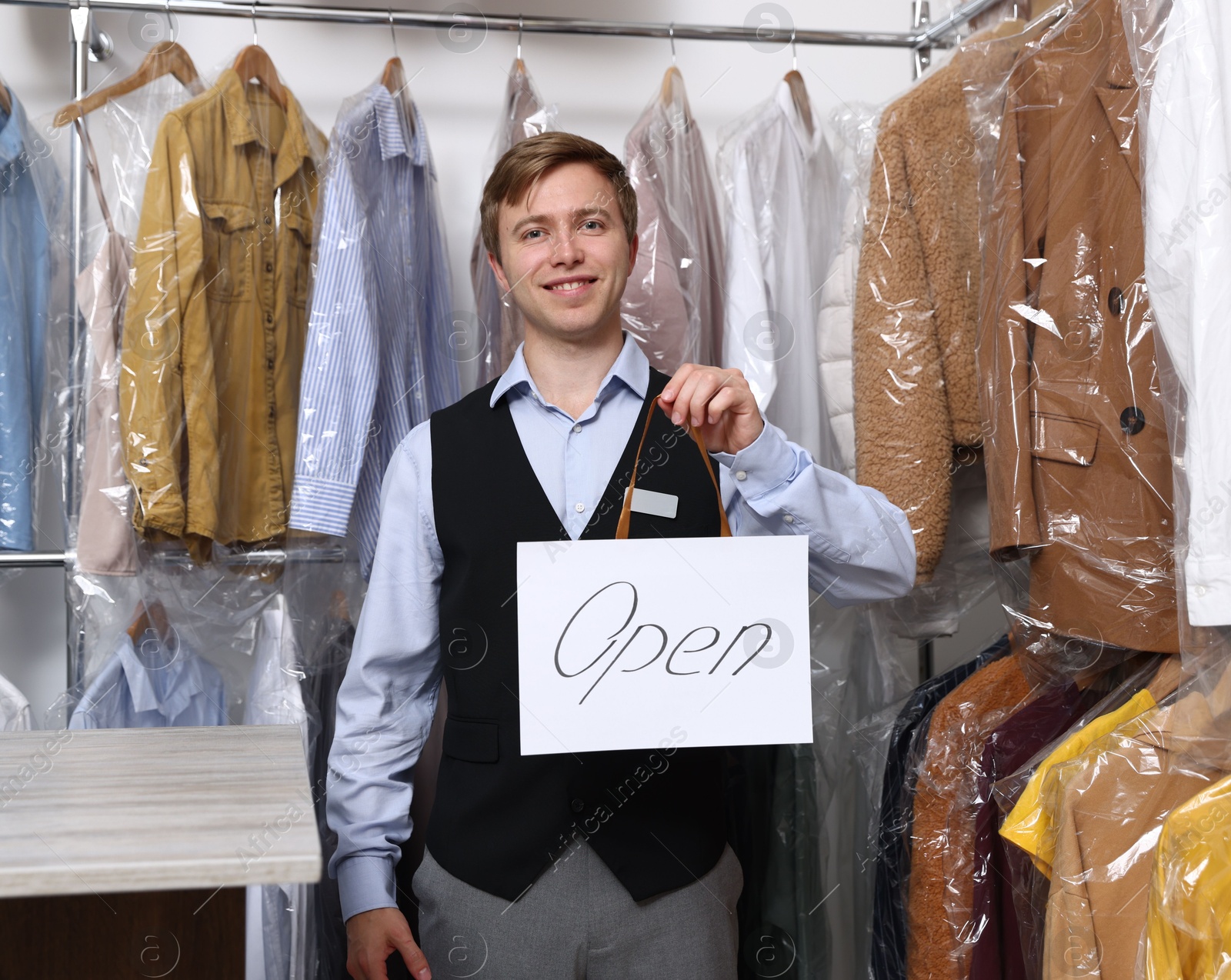 Photo of Dry-cleaning service. Happy worker holding Open sign at near rack with clothes indoors