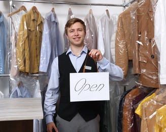 Photo of Dry-cleaning service. Happy worker holding Open sign at near rack with clothes indoors
