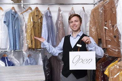Photo of Dry-cleaning service. Happy worker holding Open sign at near rack with clothes indoors