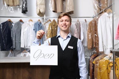 Photo of Dry-cleaning service. Happy worker holding Open sign at near rack with clothes indoors