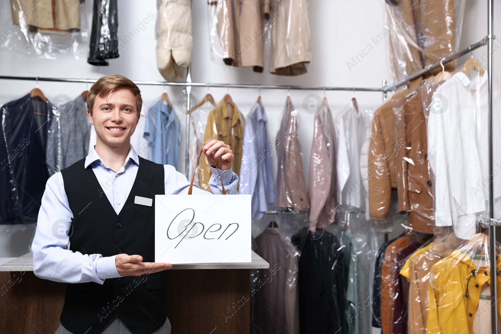 Photo of Dry-cleaning service. Happy worker holding Open sign at near rack with clothes indoors, space for text