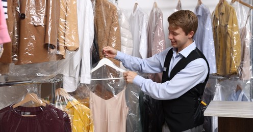 Photo of Dry-cleaning service. Happy worker choosing clothes from rack indoors