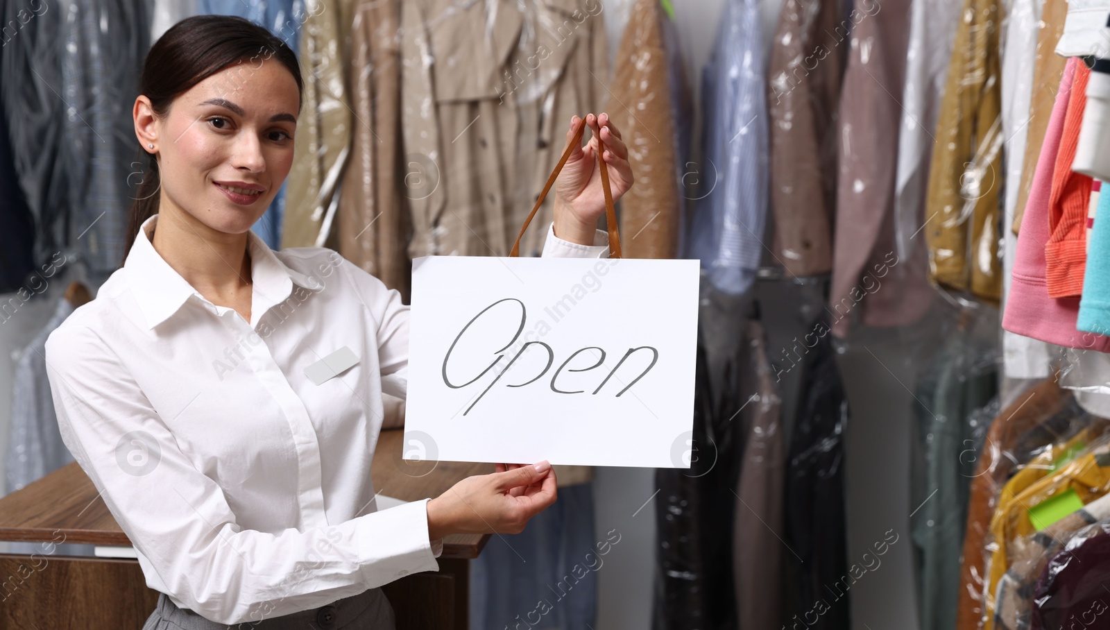 Photo of Dry-cleaning service. Happy worker holding Open sign at near rack with clothes indoors, space for text