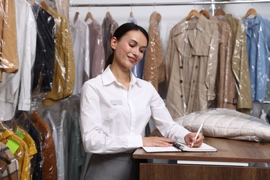 Photo of Dry-cleaning service. Happy worker writing in notebook at counter indoors