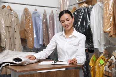 Photo of Dry-cleaning service. Happy worker writing in notebook at counter indoors
