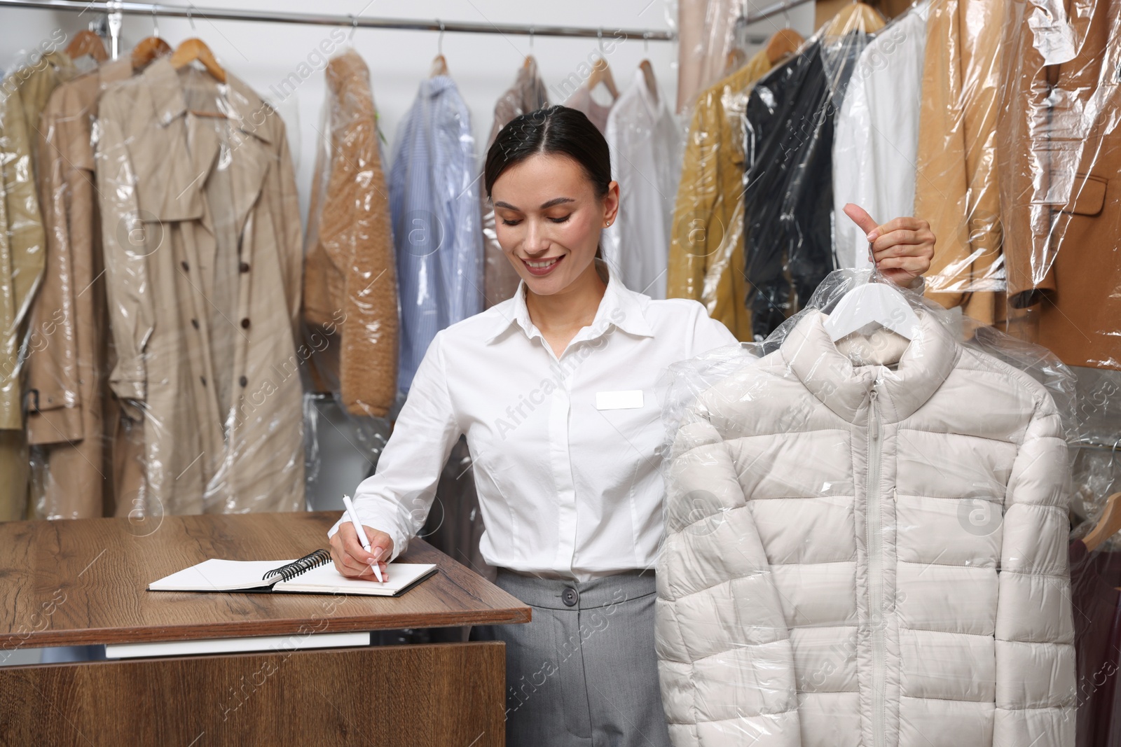 Photo of Dry-cleaning service. Happy worker holding coat in plastic bag and writing in notebook at counter indoors