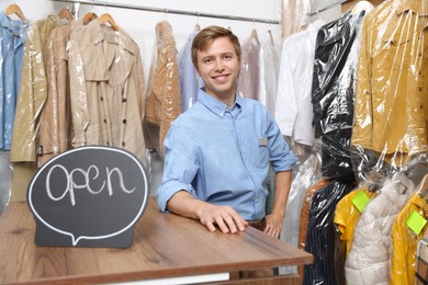 Photo of Dry-cleaning service. Happy worker with Open sign at counter indoors