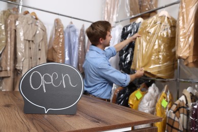 Photo of Dry-cleaning service. Worker choosing clothes from rack, focus on Open sign