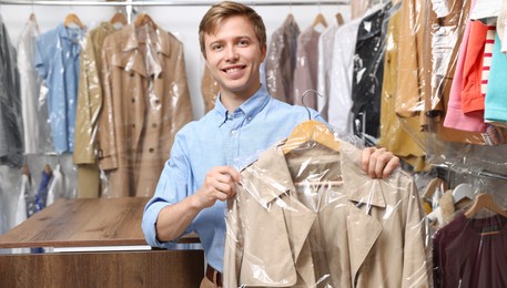 Photo of Dry-cleaning service. Happy worker holding coat in plastic bag indoors