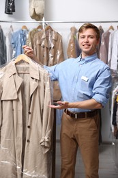 Photo of Dry-cleaning service. Happy worker holding coat in plastic bag indoors