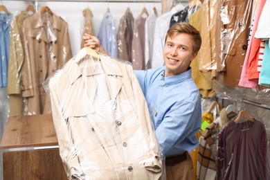 Photo of Dry-cleaning service. Happy worker holding coat in plastic bag indoors