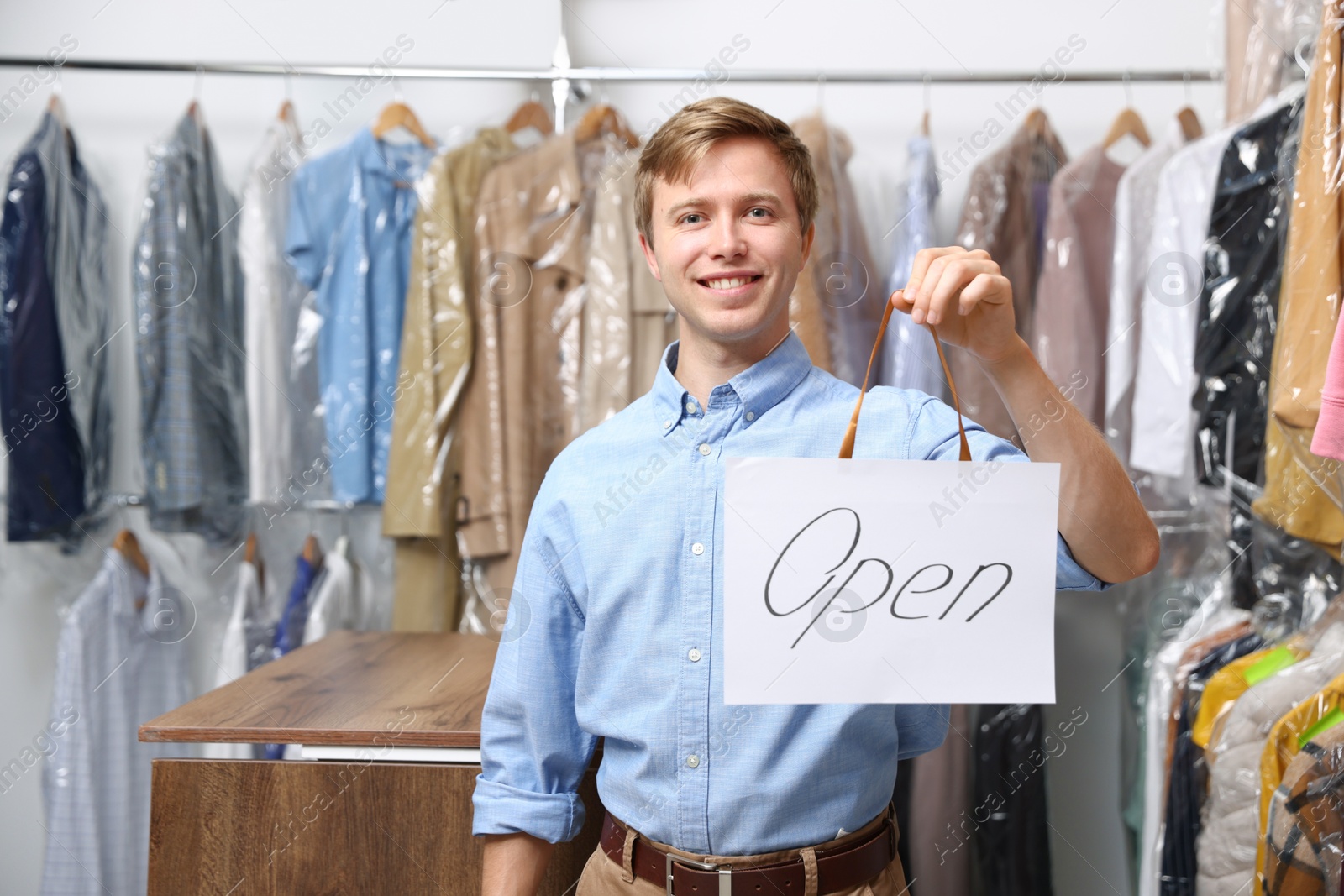 Photo of Dry-cleaning service. Happy worker holding Open sign at near rack with clothes indoors