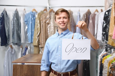 Photo of Dry-cleaning service. Happy worker holding Open sign at near rack with clothes indoors