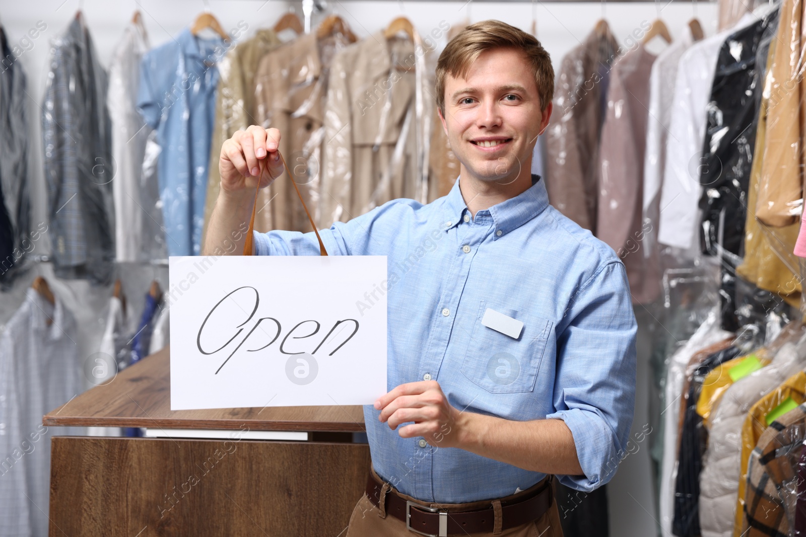 Photo of Dry-cleaning service. Happy worker holding Open sign at near rack with clothes indoors