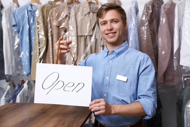 Photo of Dry-cleaning service. Happy worker holding Open sign at near rack with clothes indoors